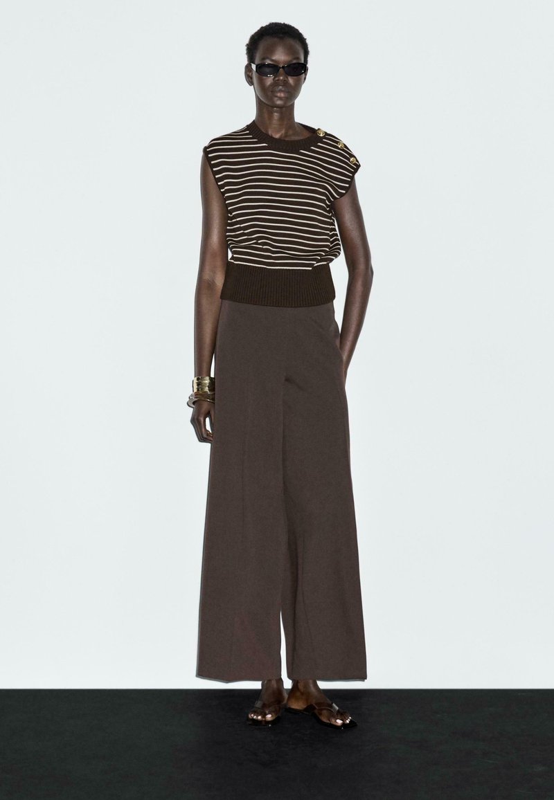 Top a righe marrone e crema in maglia con dettagli sulle spalle, abbinato a pantaloni larghi marroni. Indossato con occhiali da sole neri e sandali piatta.