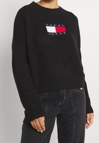 Person i en svart ribbad tröja med en röd, vit och blå Tommy Jeans-logotyp och mörka byxor med vit sömnad.