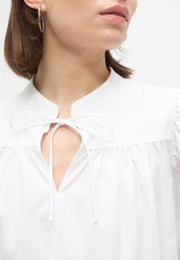 Γυναίκα με λευκό μπλουζάκι με λαιμόκοψη-κλειδαρότρυπα δεμένη με λεπτό λευκό κορδόνι και ασημένιο κρίκο ως σκουλαρίκι, στραμμένη λίγο προς τα δεξιά.