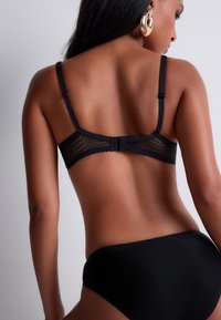 Soutien-gorge en dentelle noire avec dos en dentelle, caractéristiques des bretelles ajustables, des motifs géométriques subtils et une fermeture crochet et œil. Tissu noir lisse sur le bas.