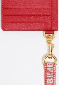 Portafoglio in pelle rossa con quattro slot per carte, superficie testurizzata, clip in metallo dorato e una cinghia intrecciata con accenti di testo crema.