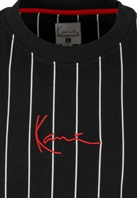 Svart T-shirt med vita vertikala ränder, med en röd broderad logotyp och en rundad halsringning. Invändig trim är röd. Storlek L.