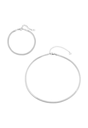 Erhobener männlicher Arm mit geballter Faust, trägt ein dünnes silbernes Armband und ein weißes ärmelloses Hemd vor einfarbigem Hintergrund.