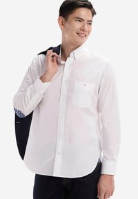 Chemise blanche à manches longues avec boutonnage, dotée d'une poche poitrine, d'un ourlet arrondi et de motifs sur les poignets intérieurs. Écharpe drapée sur l'épaule.