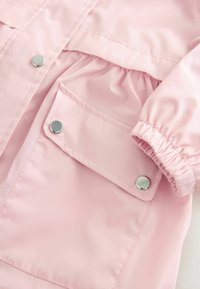 Giacca impermeabile rosa chiaro con polsini elastici, vita arricciata e bottoni a pressione argentati. Presenta una tasca frontale per la conservazione.