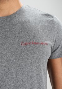 Šedé bavlněné tričko s kulatým výstřihem, na levé hrudi zdobeno červeným vyšitým logem "Calvin Klein Jeans".
