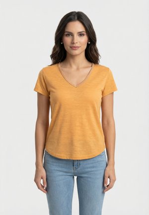 Femme aux cheveux foncés portant des boucles d'oreilles en perles, un t-shirt manches courtes à col en V jaune moutarde et un jean bleu clair, debout devant un fond uni.