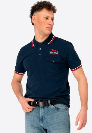 Polo azul marino con cuello, tapeta de botones y bolsillo en el pecho izquierdo a rayas rojas y blancas. Cuenta con el logo de Lonsdale en rojo.