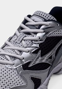 Harmaa urheilukenkä verkkokuosilla, pehmustetulla kielen alueella ja hopeanvärisillä koristeilla. Varustettu litteillä nauhoilla ja kuvioidulla pohjalla parantamaan pitoa.