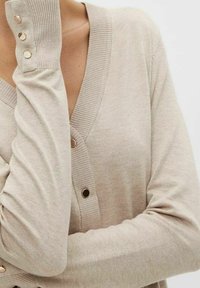Beige v-ringad cardigan med ribbad mudd och knappar i guldton. Tyget verkar mjukt med en slät textur.