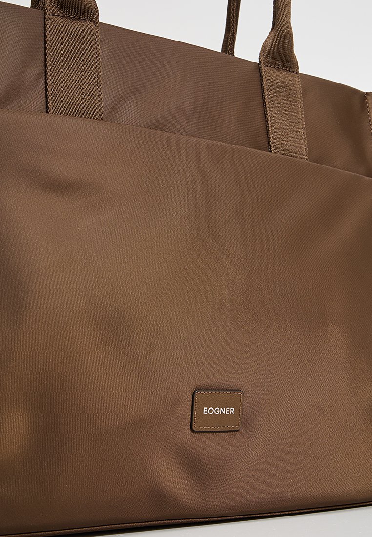 Braune Textiltasche mit robusten Trägern, ausgestattet mit einer Fronttasche und einem rechteckigen Logopatch an der Unterseite. Glatte Textur.