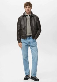 Chaqueta marrón de cuero sintético con dos bolsillos delanteros, camisa gris, vaqueros azul claro y zapatos negros. Hombre de pie contra un fondo sencillo.