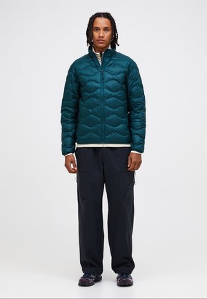 Junger Mann mit geflochtenem Haar, der eine dunkelgrüne, gesteppte Jacke, schwarze Hose und schwarze Sneakers trägt und vor einem schlichten, hellen Hintergrund steht.