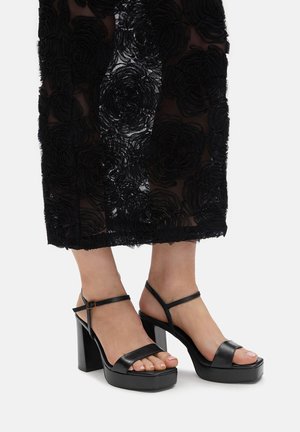 Czarne sandały na platformie z szerokimi paskami i zapięciem wokół kostki. Kawałkowy design obcasa. Noszone z czarnym materiałem w różany wzór.