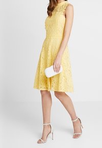 Žluté krajkové šaty s přiléhavým živůtkem a rozšířenou sukní. Průhledné lemování výstřihu, drží bílou spojku a má na sobě světlé páskové sandály.