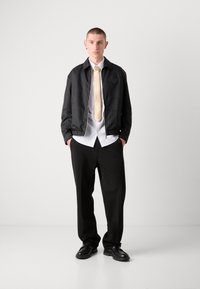 Veste bomber noire texturée avec zip, chemise blanche et cravate beige. Associée à un pantalon noir ample et des chaussures noires. Style minimaliste.
