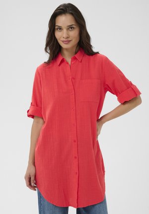 Femme aux cheveux foncés portant une chemise rouge vif à boutons, manches retroussées, debout avec une main sur la hanche, sur un fond uni.