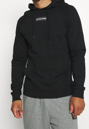 Man draagt een zwarte Jack & Jones hoodie en grijze joggingbroek, staat met ontspannen handen langs zijn zij.