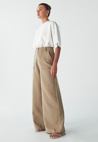 T-shirt bianco oversize con maniche scese, abbinato a pantaloni beige a gamba larga con tasche frontali e chiusura con bottone. Sandali semplici.