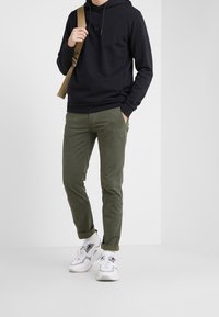 Sudadera negra con capucha y bolsillo canguro, pantalones verdes olivo ajustados y zapatillas blancas con detalles en gris, de pie sobre un fondo neutro.