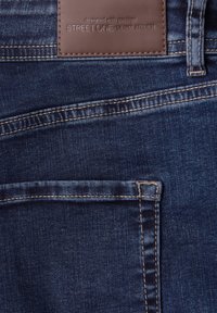 Jean denim bleu foncé avec coutures visibles sur la poche arrière et une étiquette de marque en cuir marron portant l'inscription "STREET ONE - DENIM ATELIER".