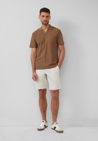 Brązowa koszula z krótkim rękawem o teksturowanym splotem i kieszonką na piersi, w połączeniu z białymi szortami oraz casualowymi trampkami z czarnymi akcentami.