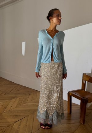 Mujer de pie en el interior, vestida con un cárdigan azul claro abotonado y una falda larga dorada y brillante con borde de encaje, junto a una silla de madera.