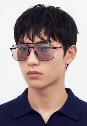 Jeune homme aux cheveux noirs courts portant de grandes lunettes de soleil carrées teintées avec des verres à motifs et une chemise bleu foncé à col.