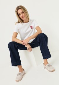Mujer con cabello rubio que lleva una camiseta blanca con la palabra "LOVE", vaqueros azul oscuro y zapatos tipo slip-on beige, sentada en un cubo blanco sobre un fondo liso.