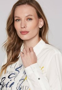Camicia bianca con bottoni, caratterizzata da un testo grafico colorato. Tessuto morbido con colletto classico e polsini dettagliati accentuati da bottoni.