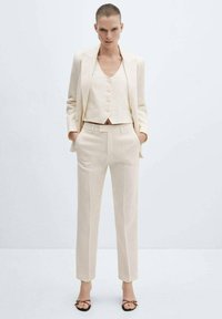 Beige three-piece suit featuring a cropped blazer, buttoned vest, and tailored pants with a smooth texture and clean lines.