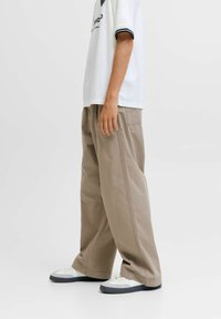 Persona che indossa una maglietta bianca con grafica, pantaloni beige larghi e sneakers bianche con suola grigia, in piedi contro uno sfondo neutro.