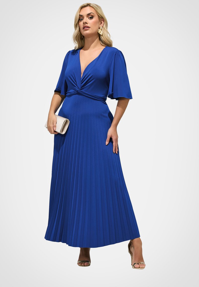 Frau in einem tiefblauen, plissierten Maxikleid mit Ellbogen-langen Ärmeln, hält eine beige Clutch, trägt goldene Absätze und Ohrringe.