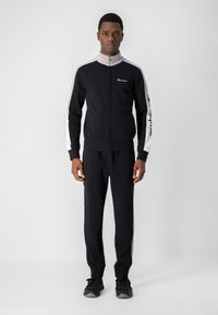Tuta nera in tessuto morbido con giacca con zip caratterizzata da accenti grigi e logo, abbinata a pantaloni coordinati, che mette in mostra un design sportivo.