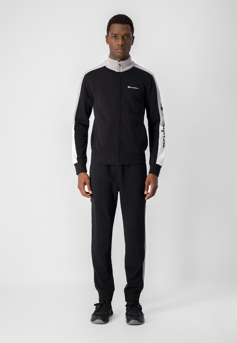 Tuta nera in tessuto morbido con giacca con zip caratterizzata da accenti grigi e logo, abbinata a pantaloni coordinati, che mette in mostra un design sportivo.