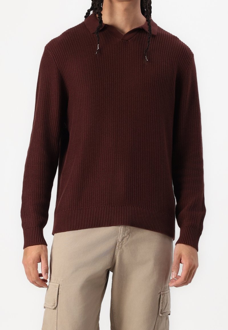 Homme aux cheveux tressés, portant un pull en maille texturée marron foncé à manches longues et un pantalon cargo beige, debout devant un fond uni.