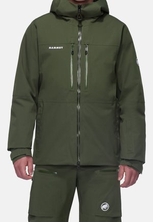 Homme portant une veste et un pantalon Mammut vert olive avec plusieurs poches zippées et des écussons logo sur la poitrine et la jambe.