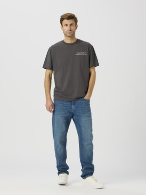 Graues Baumwoll-T-Shirt mit kurzen Ärmeln und einem Aufdruck auf der linken Brust, kombiniert mit blauen Jeans und weißen Turnschuhen.