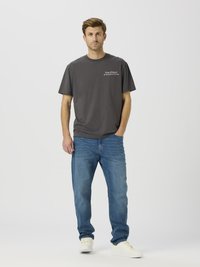 Hall puuvillane T-särk lühikeste varrukatega ja vasakul rinnal trükitud mustriga, kombineeritud siniste teksapükste ja valgete tossudega.