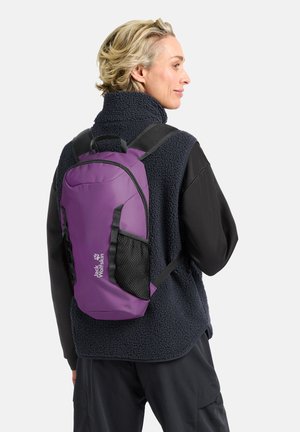 Persoon die een marineblauw fleecevest en zwarte broek draagt met een paarse Jack Wolfskin-rugzak waarin zijvakken van mesh zichtbaar zijn.