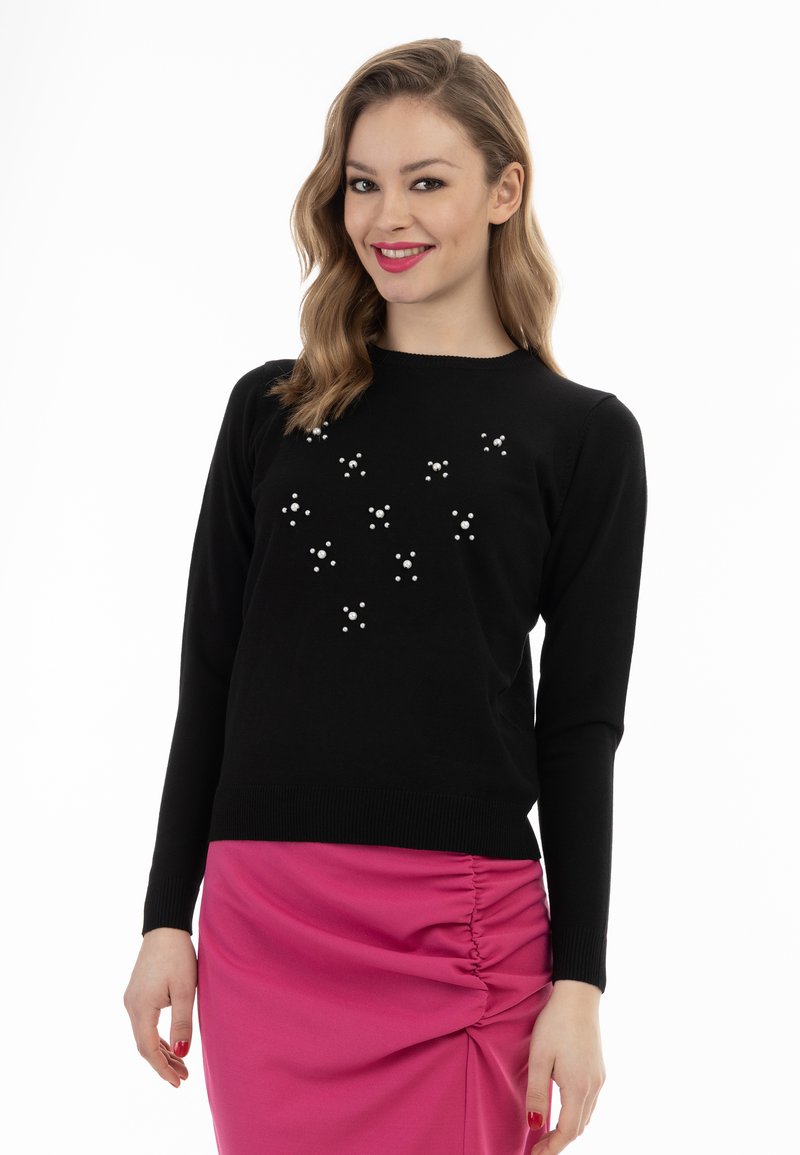 Schwarzer Strickpullover mit tiefem Rundhalsausschnitt, der mit einem Muster aus weißen Perlen verziert ist. Kombiniert mit einem rosa gerafften Rock. Lange Ärmel und gerippte Bündchen.