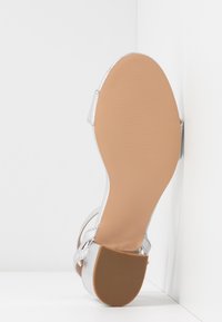 Sandale à talons hauts argentée avec un dessus lisse, une bride à boucle et une semelle extérieure en cuir texturé marron clair. Présente un design classique à bout ouvert.