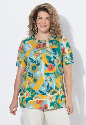 Frau mit lockigem Haar lächelt, trägt ein kurzärmeliges Oberteil mit Blumenmuster in Türkis, Gelb, Rot und Weiß, kombiniert mit hellen Hosen.