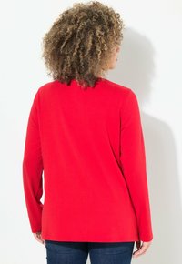 Rode longsleeve shirt gemaakt van zachte stof, met een relaxte pasvorm en ronde halslijn, gedragen met marineblauwe jeans, bekeken van achteren.