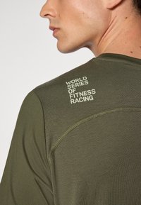 Oliven grønn idretts skjorte med en glatt tekstur, med et trykt logo "WORLD SERIES OF FITNESS RACING" på skulderen.
