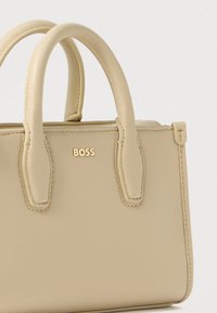 Μπεζ δερμάτινη τσάντα χειρός με δύο στρογγυλεμένες λαβές. Διαθέτει ανάγλυφο λογότυπο "BOSS" σε χρυσό. Λείο υλικό, δομημένο σχήμα, μινιμαλιστικός σχεδιασμός.