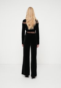 Μαύρο βελούδινο σύνολο δύο τεμαχίων με μπλούζα μακρυμάνικη με διάφανες επιφάνειες και παντελόνια φαρδιάς γραμμής, σε συνδυασμό με ψηλοτάκουνα παπούτσια.