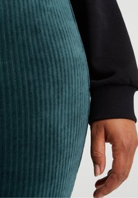 Turkusowa tkanina w prążki o gładkiej fakturze, połączona z czarnym, dzianinowym rękawem; przedstawia zbliżenie dłoni opierającej się na tkaninie.