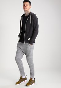 Dunkelgrauer Reißverschluss-Hoodie mit weißem Kordelzug, schwarzes T-Shirt, hellgraue Jogginghose und dunkelolivgrüne Sneaker mit weißen Akzenten.
