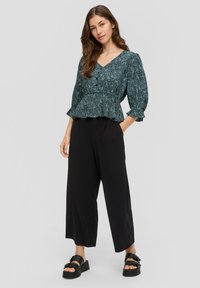 Grüne Blumen-Peplum-Bluse mit V-Ausschnitt und Knopfleiste, kombiniert mit schwarzen weit geschnittenen Hosen und schwarzen Plateausandalen.